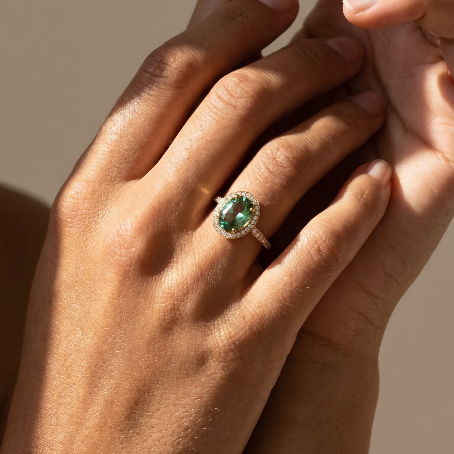 Mink Green oval cut tourmaline Close-up of two hands with a ring featuring an emerald stone on a neutral background