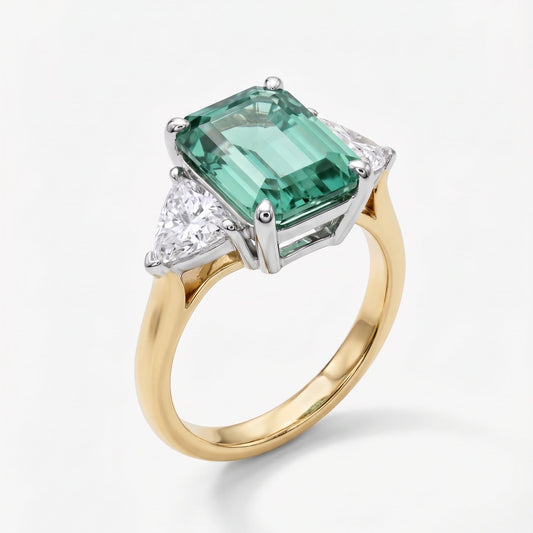 A yellow gold ring with a large emerald-cut green gemstone and small clear accent stones in a silver-toned setting, shown on a light gray background.