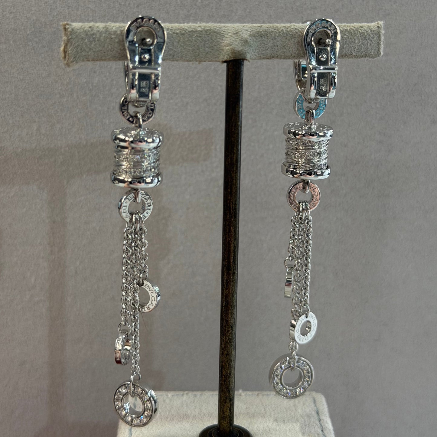 A pair of silver-toned chain earrings with textured spacers and clear stones, displayed on a black horizontal bar mounted on a light gray fabric stand.