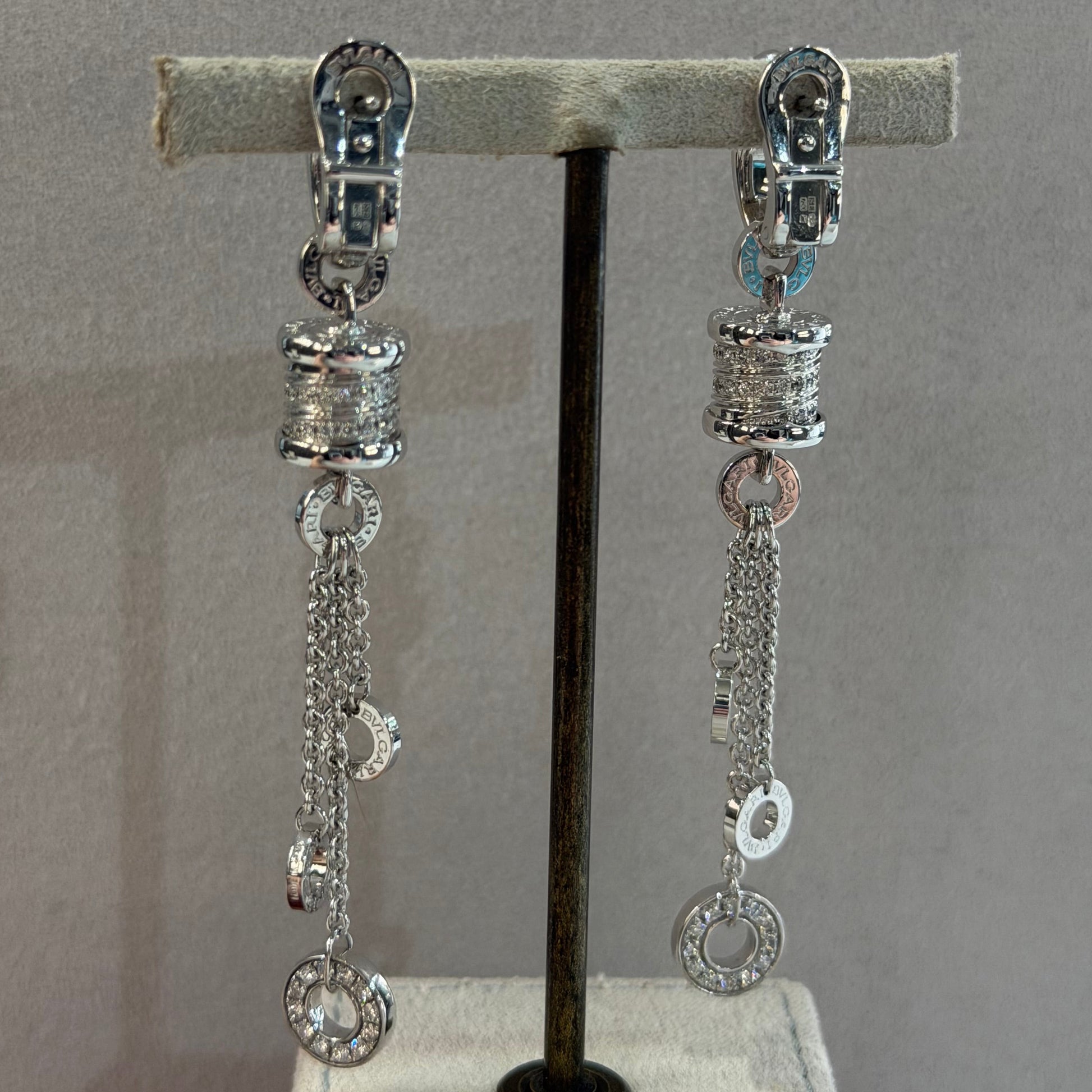 A pair of silver-toned chain earrings with textured spacers and clear stones, displayed on a black horizontal bar mounted on a light gray fabric stand.