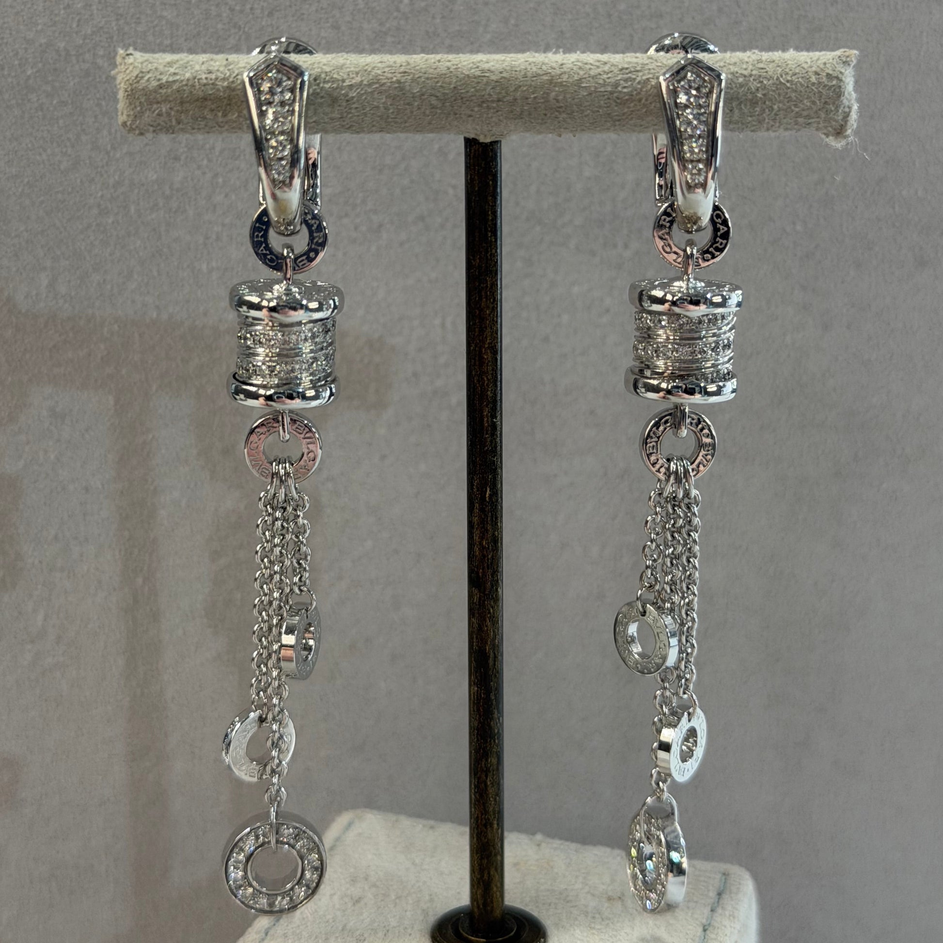 A pair of silver-toned dangle earrings with clear rhinestone-encrusted beads, chain links, and oval ring charms, displayed on a gray jewelry stand.