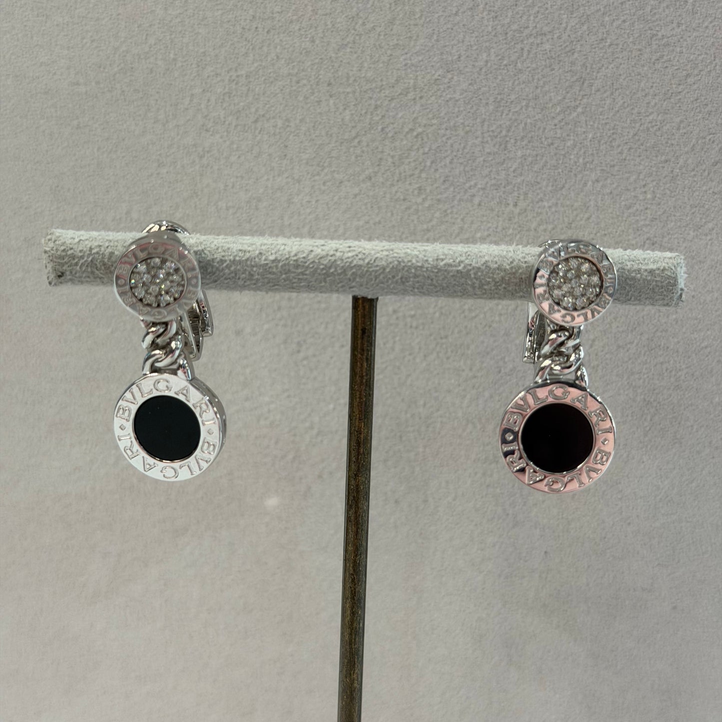 A pair of silver-toned hoop earrings with clear pavé-set round stones and a central black circular inlay, displayed on a gray jewelry stand.