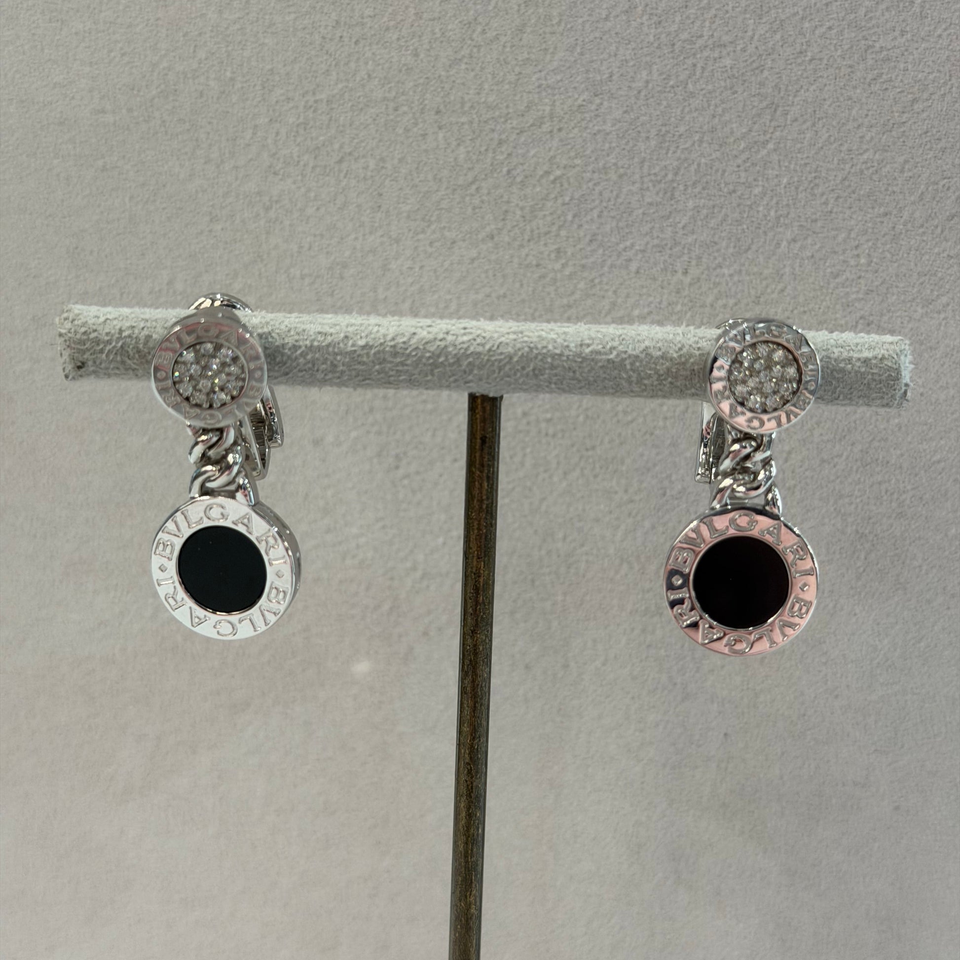 A pair of silver-toned hoop earrings with clear pavé-set round stones and a central black circular inlay, displayed on a gray jewelry stand.