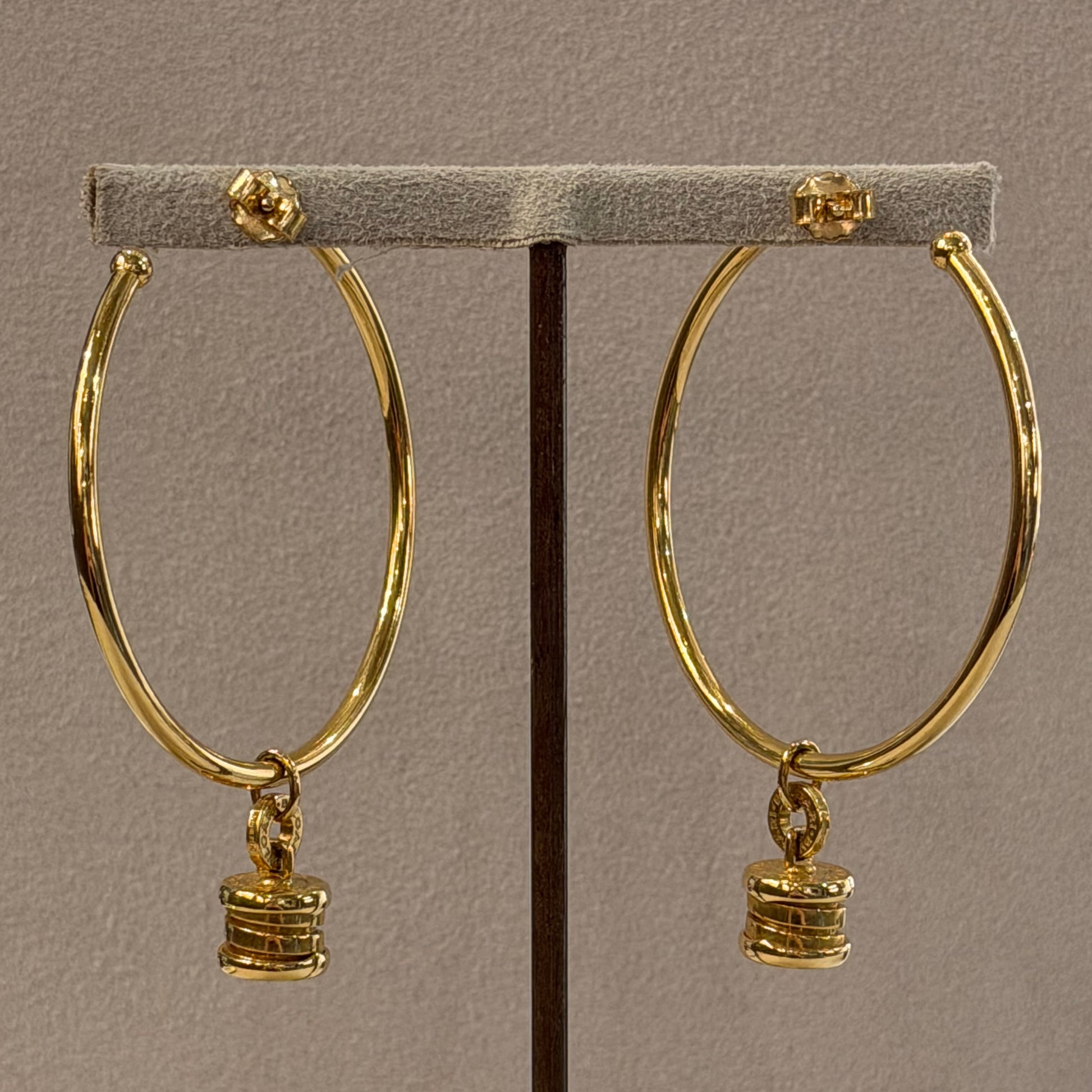A pair of gold hoop earrings with small stacked disc charms, displayed on a gray jewelry stand against a neutral background.