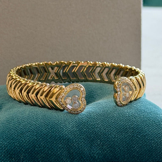 A gold-plated open cuff bracelet with heart-shaped clover motifs and pavé-set clear stones, displayed on a teal velvet surface.