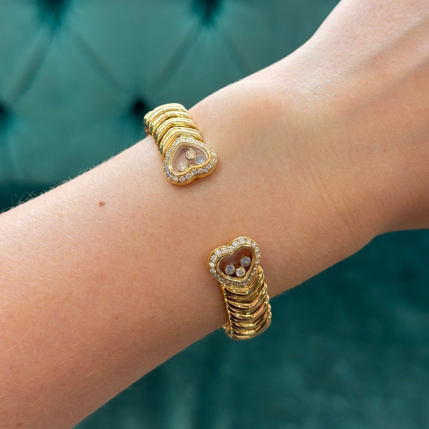 A close-up of a person's wrist wearing two gold cuff bracelets with heart-shaped crystal and bead details, set against a teal tufted background.