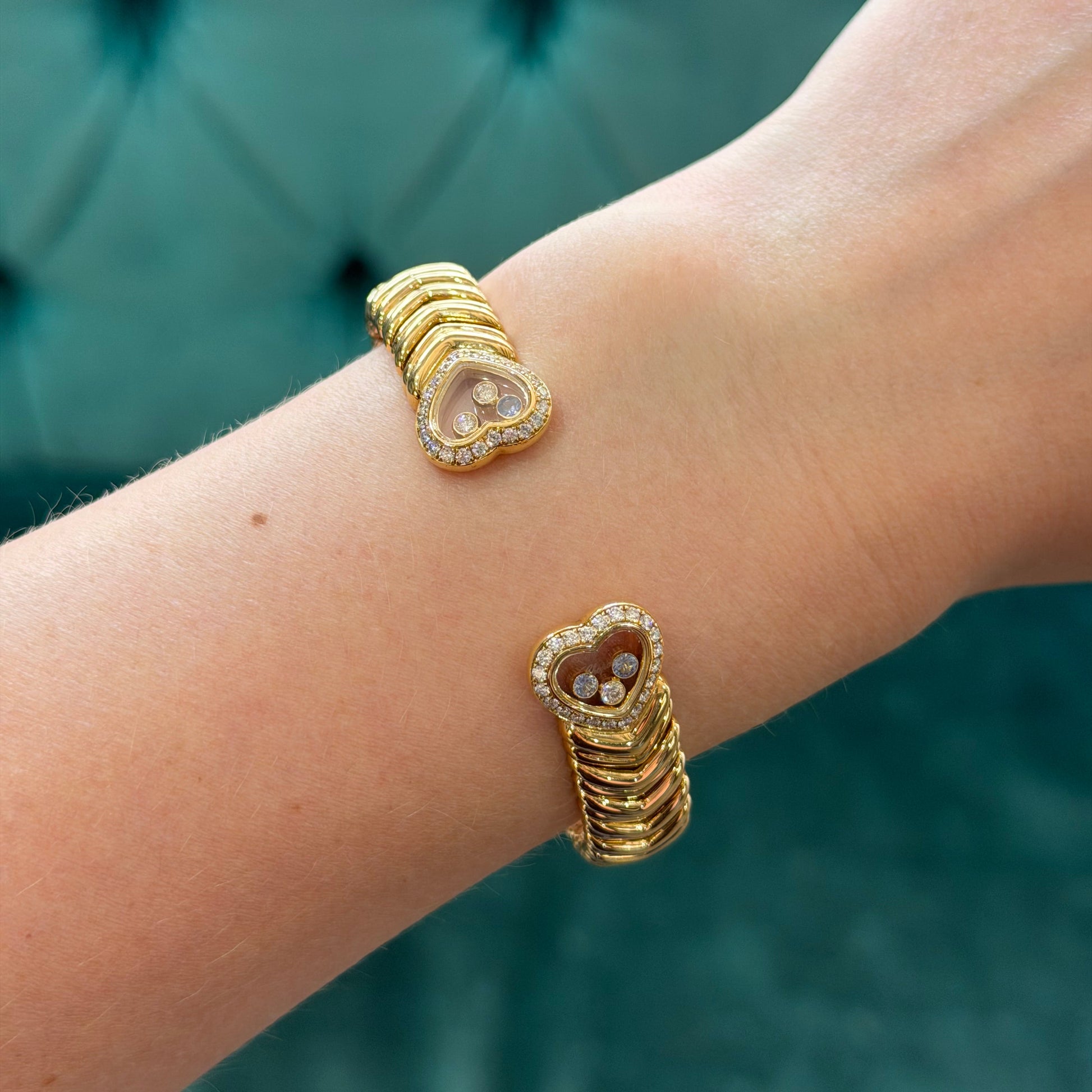 A close-up of a person's wrist wearing two gold cuff bracelets with heart-shaped crystal and bead details, set against a teal tufted background.