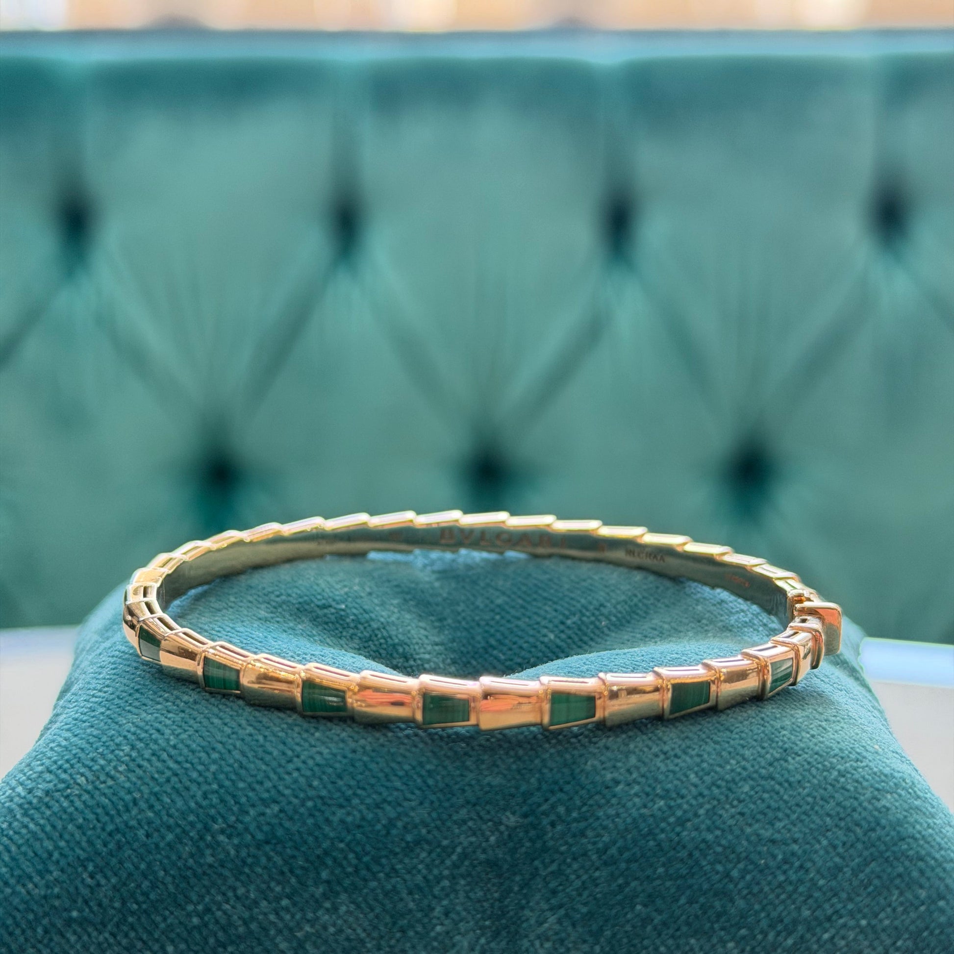 A gold bangle bracelet with rectangular green stone accents, displayed on a teal velvet cushion against a matching tufted background.