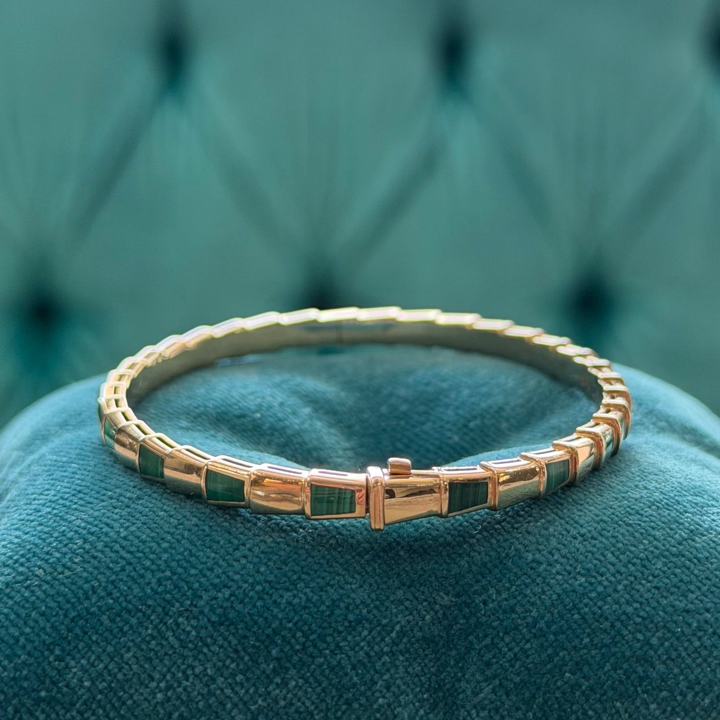A gold-plated bangle bracelet with green rectangular inlay segments, resting on a plush teal velvet cushion.