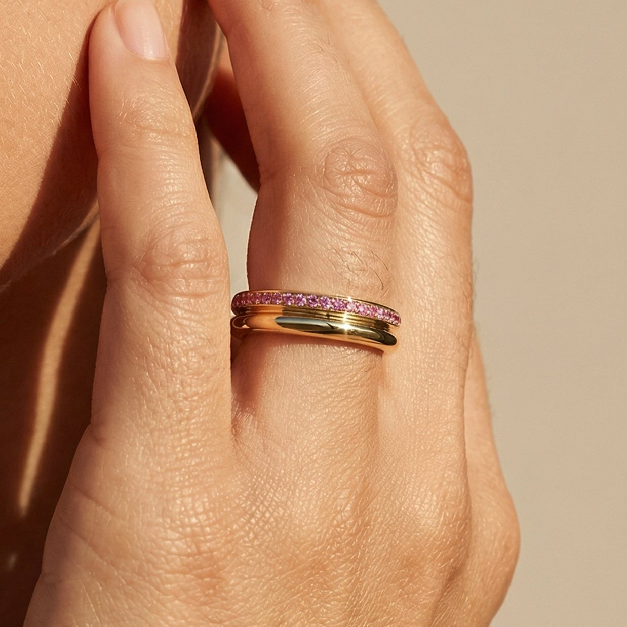 Close-up of a hand wearing a yellow gold ring with a pink sapphires against a beige background