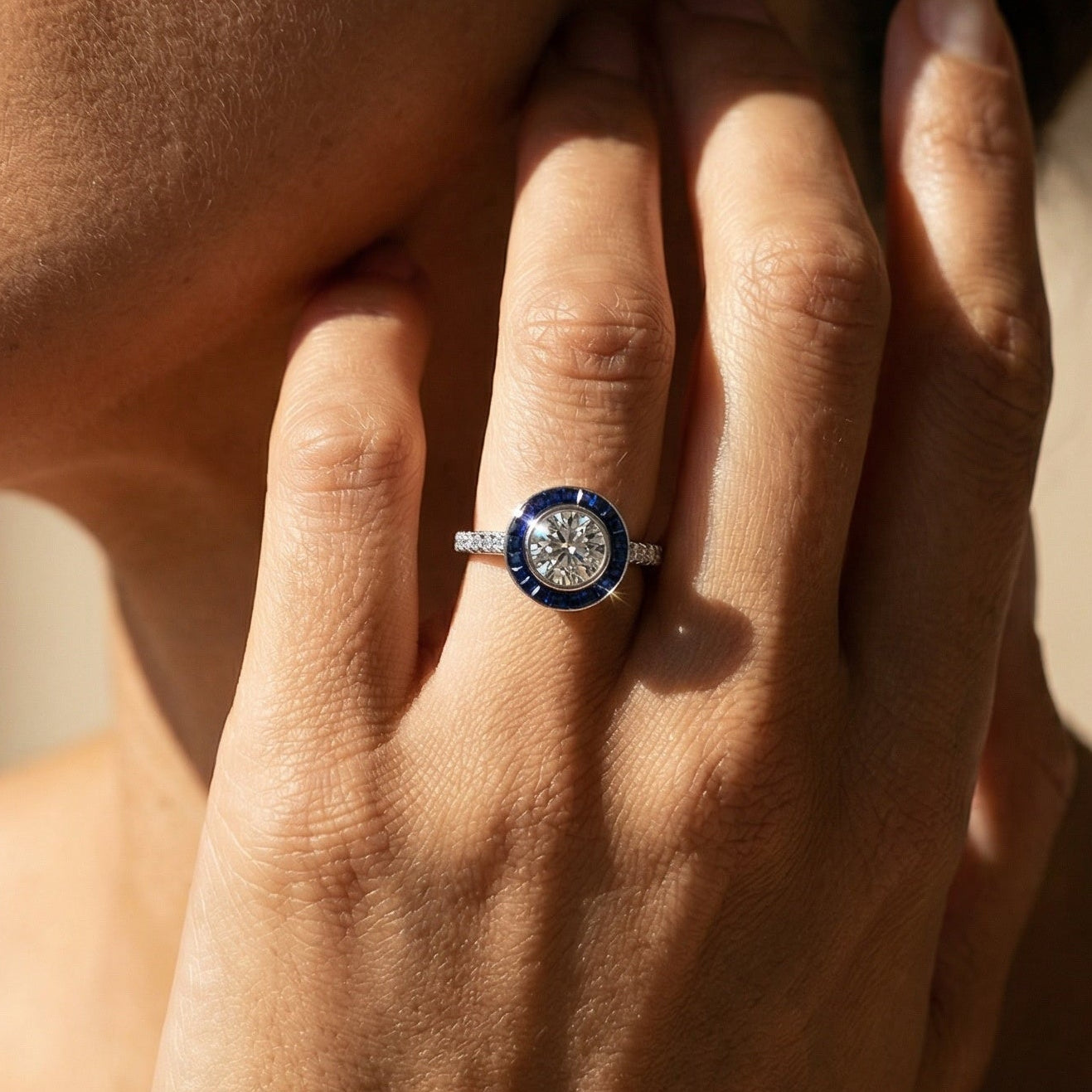 Close-up of a hand wearing a ring with a blue gemstone, touching the face.