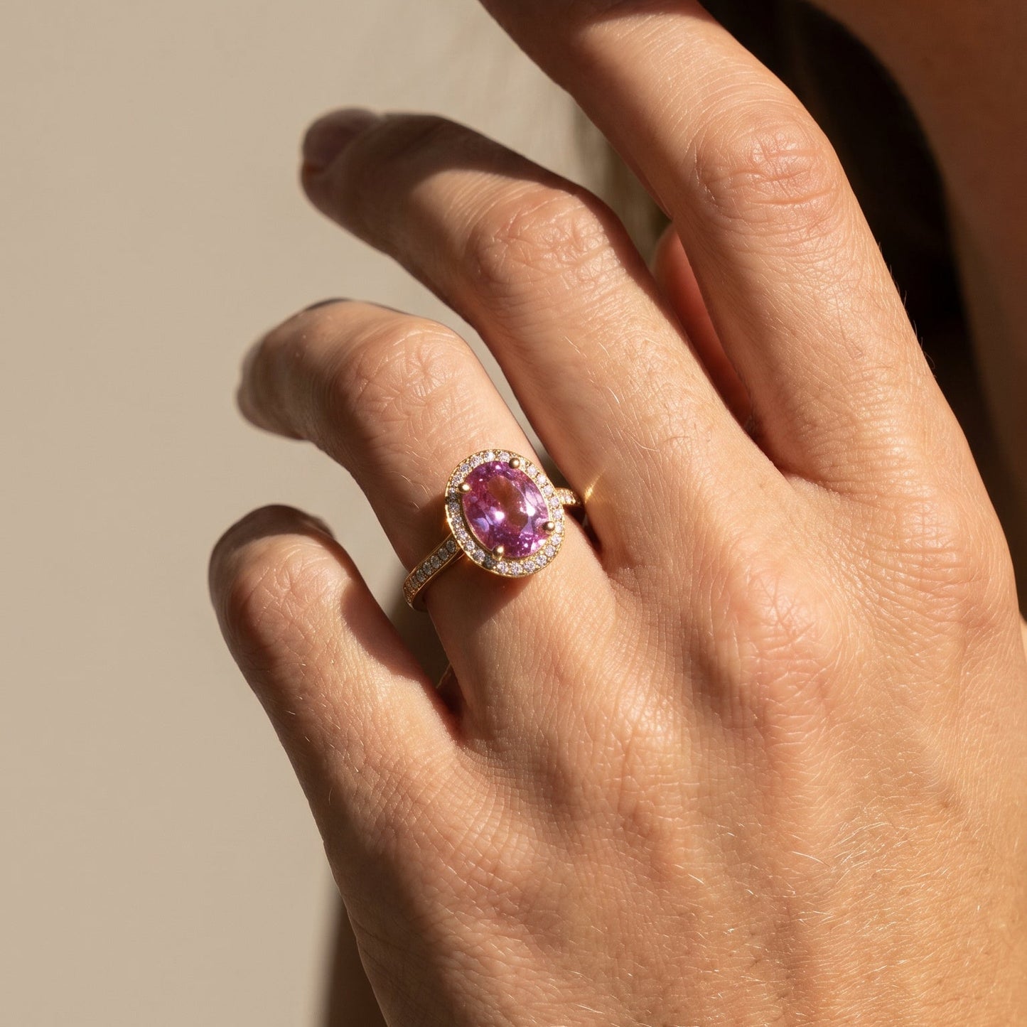 Hand wearing a ring with a pink tourmaline oval cut stone set in 18ct yellow gold  on a neutral background